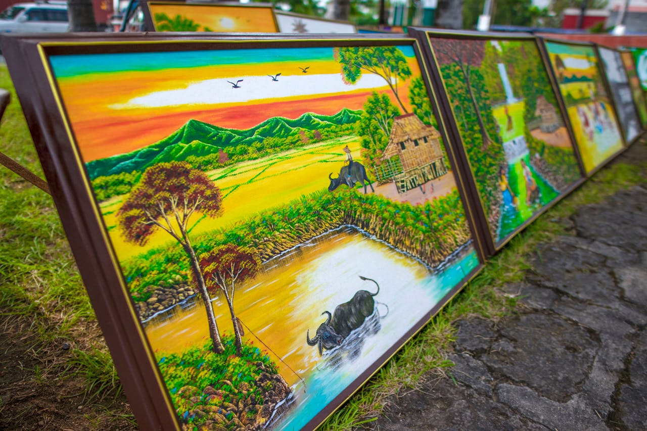 Colorful landscape paintings depicting rural scenes with mountains and buffaloes arranged outdoors.